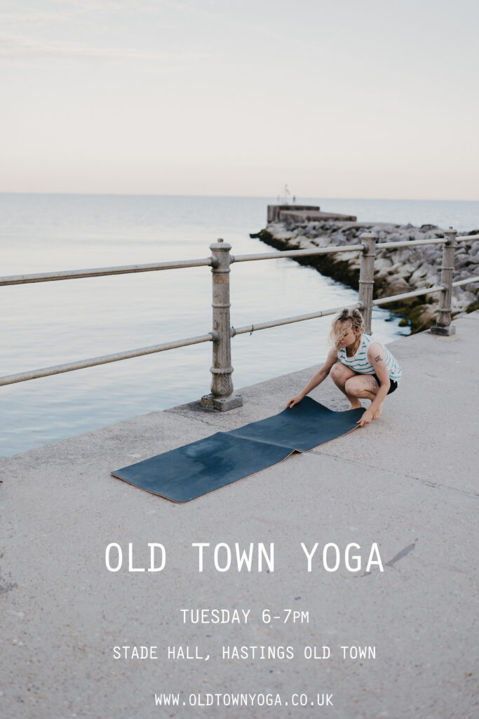 A photo of a woman laying out a yoga mat near the sea. Overlaid text reads: "Old Town Yoga. Tuesday 6-7pm. Stade Hall, Hastings Old Town. www.oldtownyoga.co.uk"