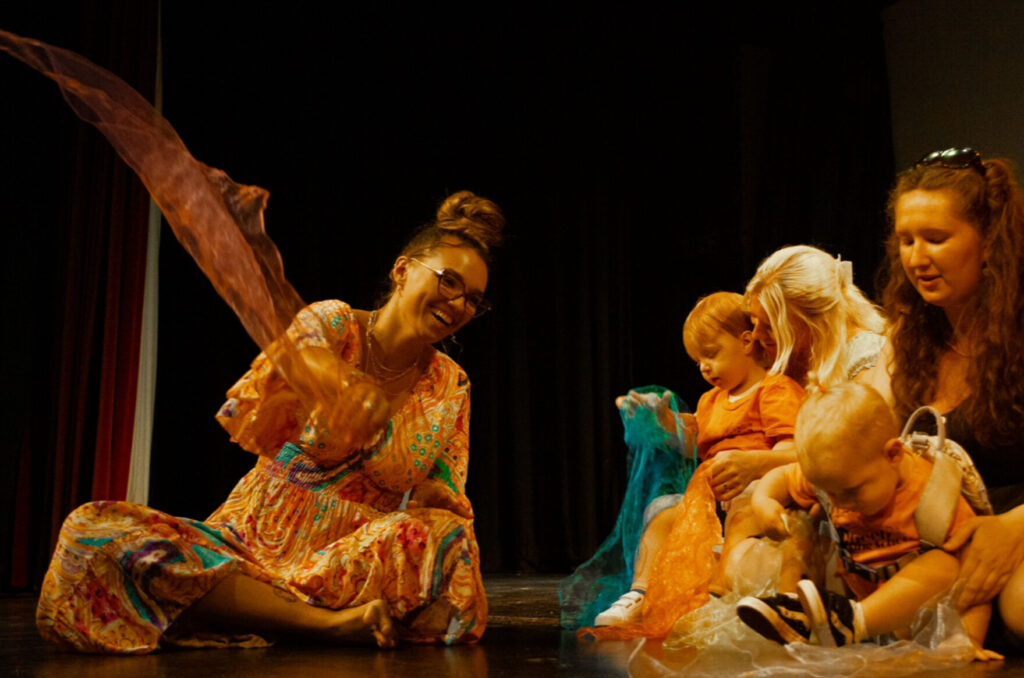A photo of two women and a baby. They are wearing bright colours, and the woman on the left is swaying some fabric.